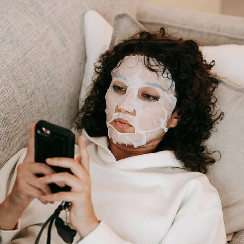 Image of woman laying on sofa, using cellphone while wearing a face mask. 
